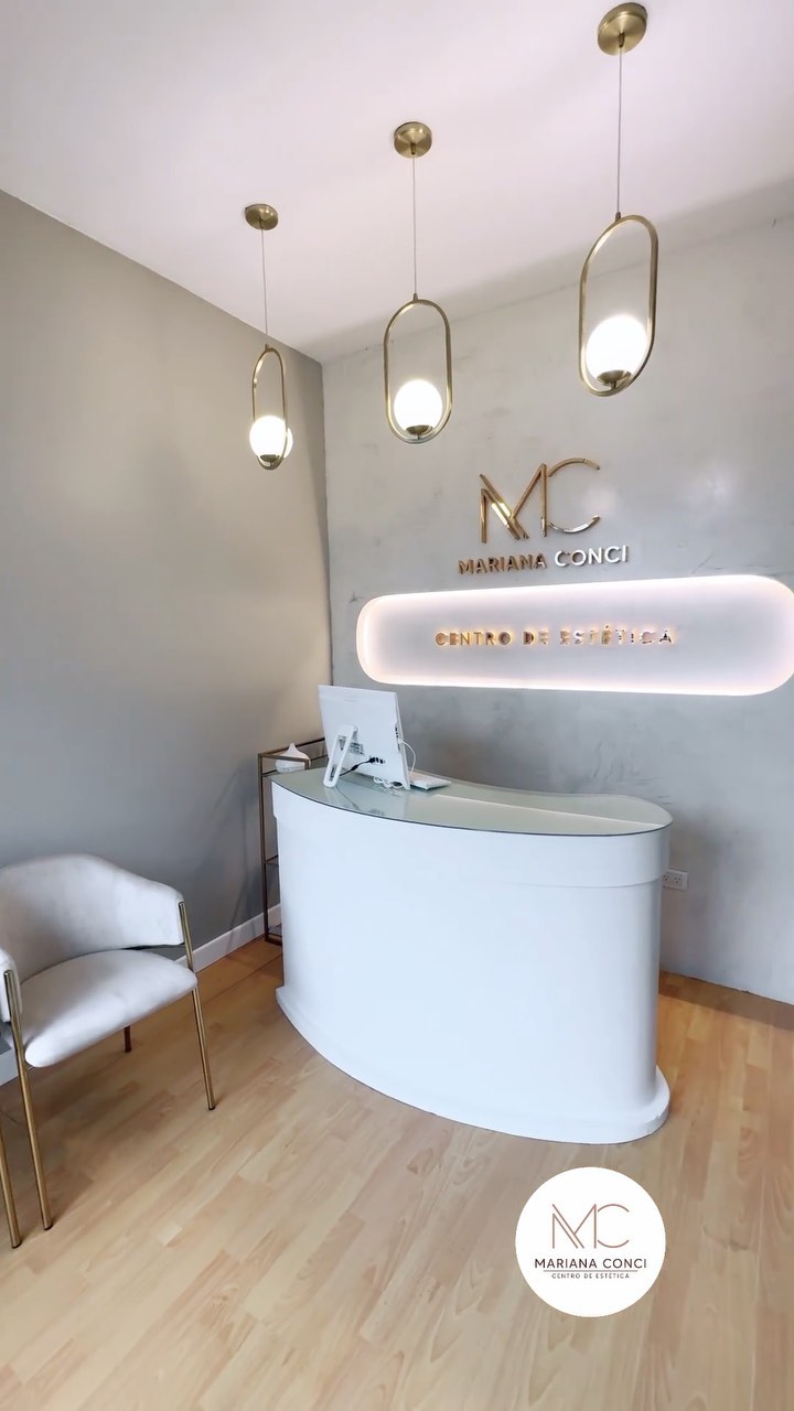 La recepción de Mariana Conci: mostrador blanco, lámparas doradas colgantes y el logo MC dorado sobre la pared gris